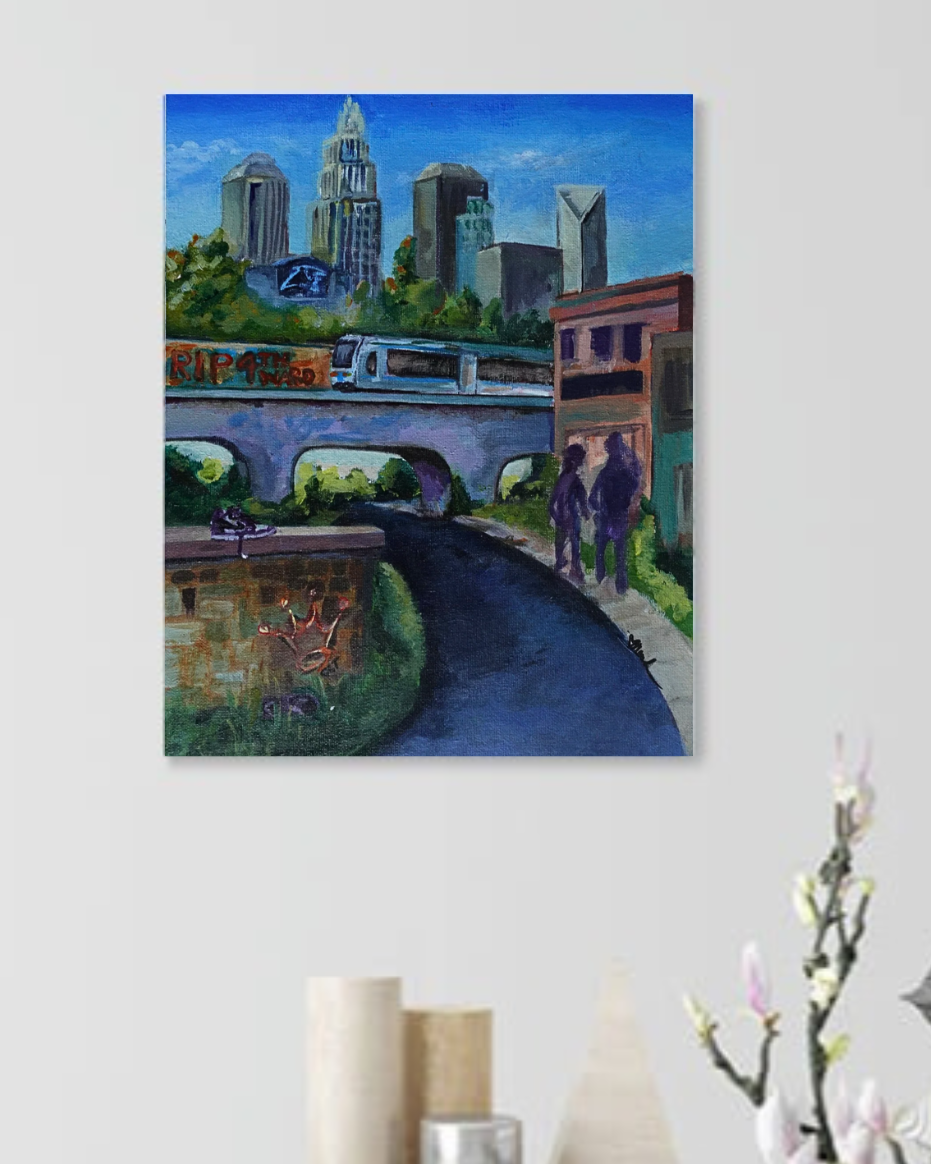 “Dear CLT” by Nesha Cornett of Love Neesh Art. A vibrant acrylic cityscape of Charlotte, North Carolina. The skyline features recognizable skyscrapers under a clear blue sky, while a light rail crosses a bridge in the midground. Below it, graffiti reading “RIP 4th Ward” is painted on a wall, referencing the gentrification of a historically Black neighborhood. In the foreground, a pair of Jordans rest on a brick ledge marked with a crown tag. Two dark silhouettes walk along a shaded path, surrounded by urban greenery and layered textures. The painting blends realism with symbolism, honoring community, memory, and change.